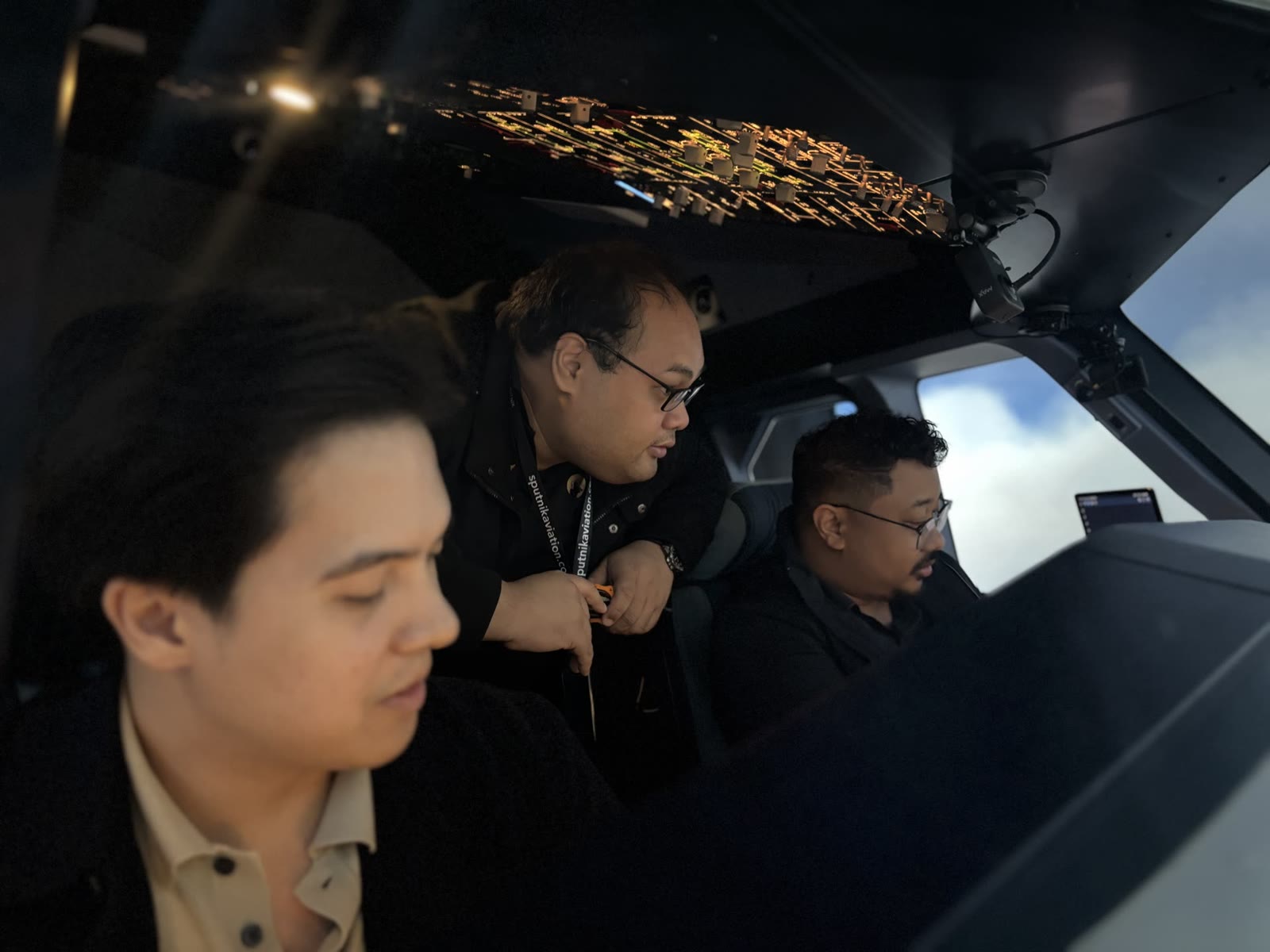 Three APOP First Officer trainees observing the A320 simulator flight deck controls together during a training session.