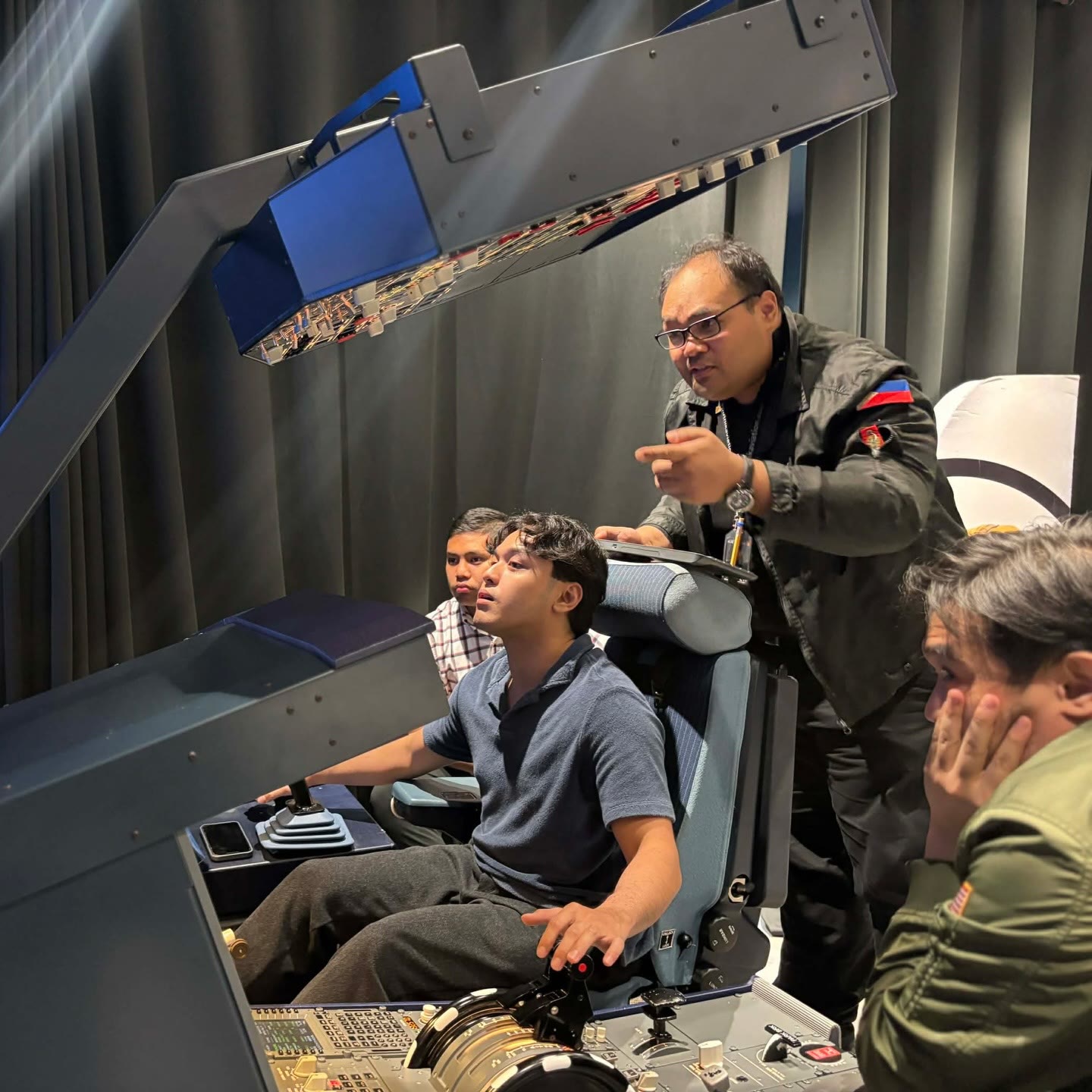An APOP First Officer instructor guiding a trainee at the full-motion A320 simulator controls, two other trainees observing.