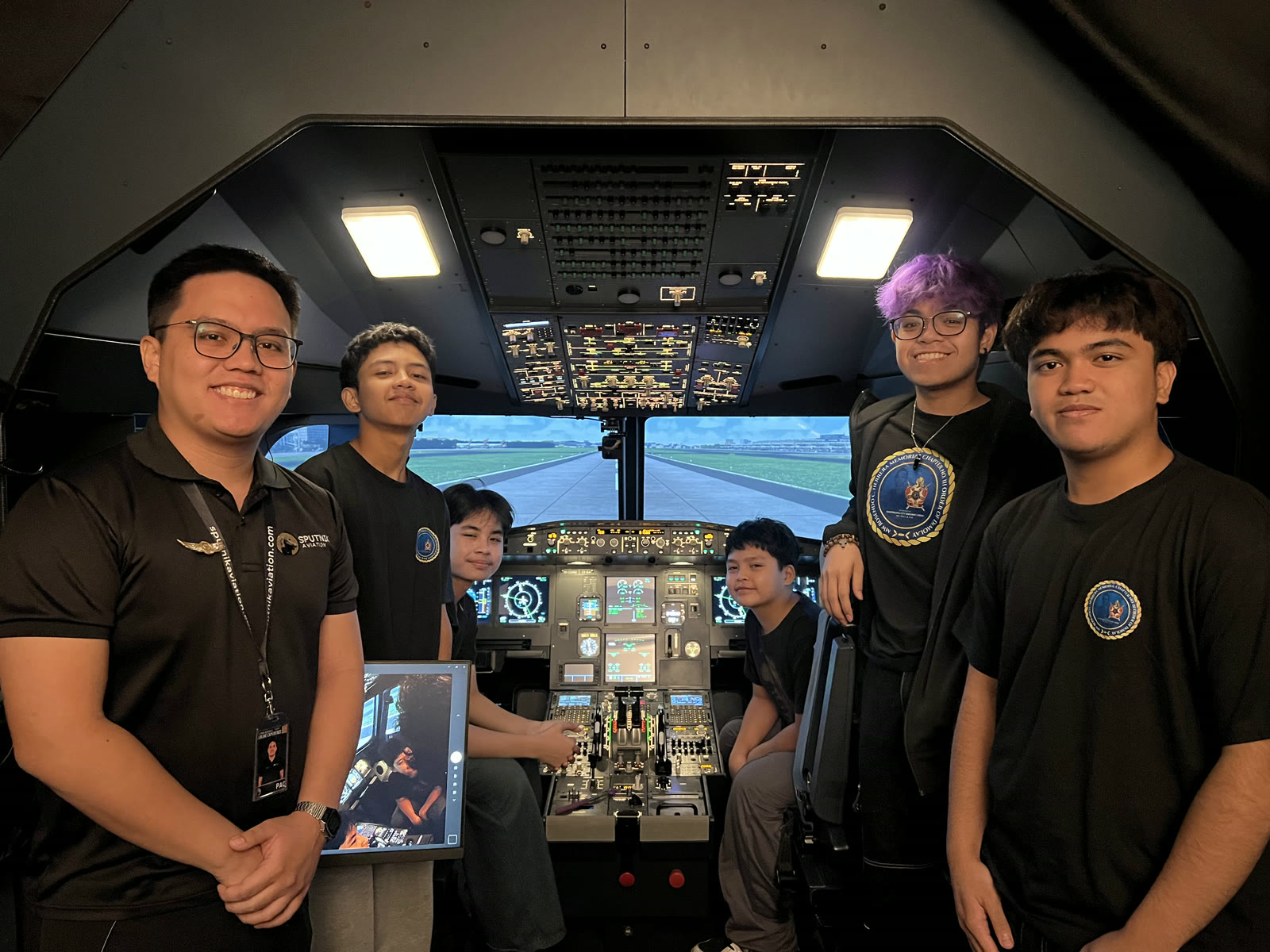 A group of six friends and staff gathered around the A320 flight deck simulator controls, smiling together with the airport runway visible on the outside visual.