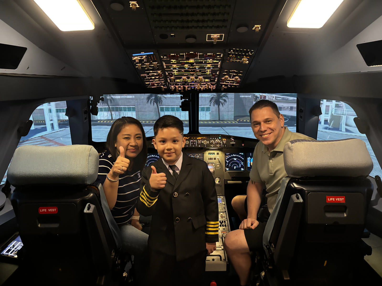 A Future Flyer participant in a captain’s uniform giving a thumbs up in the A320 flight deck simulator, flanked by his smiling parents.